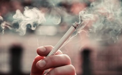 A closeup photo of fingers holding a lit cigarette with smoke wafting in the background.